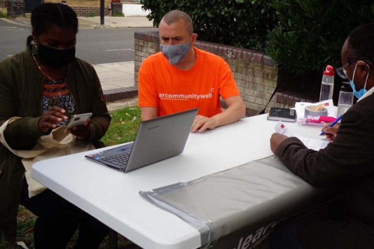 People wearing masking sat at a table outside, a man in an orange t-shirt is showing a woman how to use a laptop