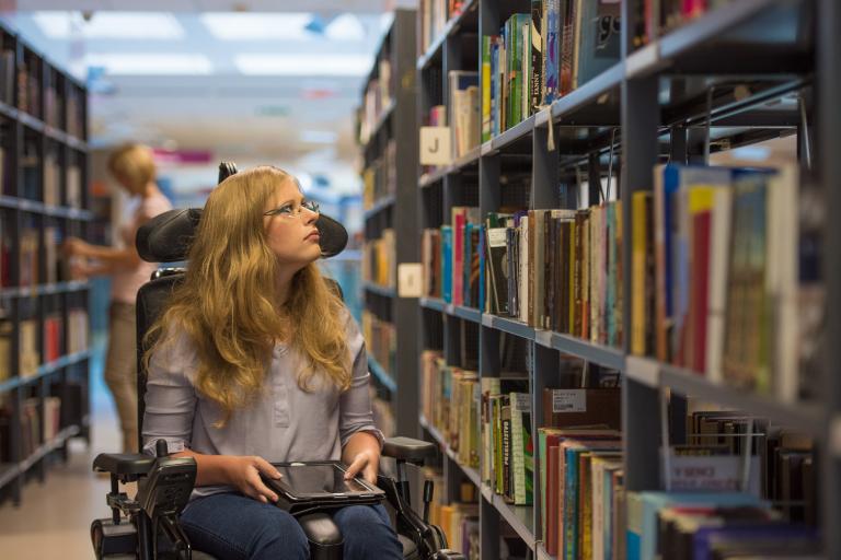 Woman in a motorised wheelchair, navigating down an aisle in a library